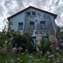 Eine bemalte Hauswand mit Blumen im Vordergrund und einem Wolkenhimmel im Hintergrund